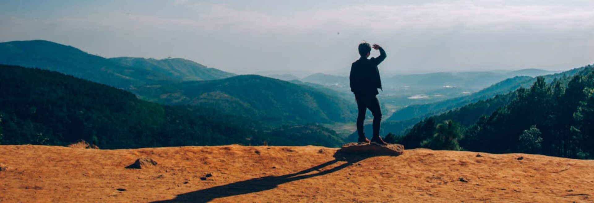 A lone traveler stands on a hilltop, gazing over a vast mountain landscape under a clear blue sky.